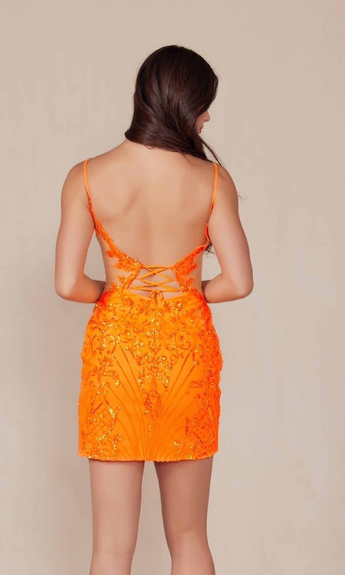 A person with long brown hair wears the Sheer-Sides Short Sequin-Print Hoco Dress D852, a bright orange sleeveless, backless party dress with lace-up details and bead embellishments, standing against a plain background.